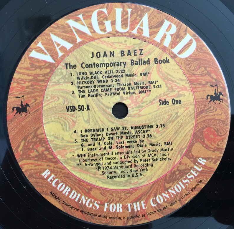 May include: A yellow and black vinyl record with the text "Vanguard" in large, bold letters. The record is titled "Joan Baez: The Contemporary Ballad Book" and features a list of songs with their titles, artists, and running times. The record is labeled "Side One" and includes the text "Recordings for the Connoisseur".