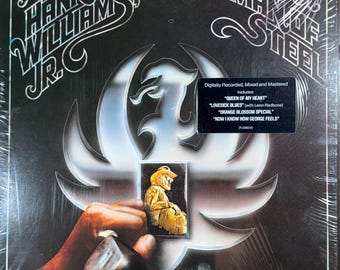 May include: A vinyl record album cover for Hank Williams Jr. titled Man of Steel. The cover features a metallic eagle emblem, a gold-toned figure, and a black sticker listing tracks including Queen of My Heart and Lovesick Blues.