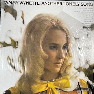 以下が含まれることがあります： タミー・ワイネットの「Another Lonely Song」のアルバムカバー。ブロンドの髪の女性が黄色のリボンを身につけています。アルバムタイトルとアーティスト名は上部にあります。アルバムはビニールスリーブに入っています。