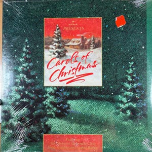 Puede incluir: Un álbum navideño de Hallmark titulado "Carols of Christmas", con una escena nevada con una casa y árboles de hoja perenne. La portada del álbum es predominantemente verde con una pancarta y texto rojos. El álbum está sellado en plástico.