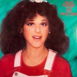 May include: Album cover featuring Gilda Radner with the text "Gilda Radner Live From New York." Radner is wearing a red shirt and white overalls. Her dark hair is styled in a voluminous, curly fashion. The background is a teal color.