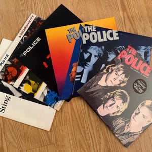 Może przedstawiać: Kolekcja płyt winylowych zespołu The Police, z okładkami albumów o różnych wzorach i kolorach. Jedna okładka jest biała z nadrukami "Sting" i "The Police". Na drewnianej powierzchni.