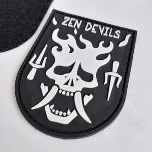 May include: Black and white patch with a skull in flames and the text "ZEN DEVILS". The skull has two large fangs and is surrounded by two weapons, a trident and a dagger.