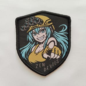 Op de afbeelding: Een schildvormige geborduurde patch met een zwarte rand, voorzien van een anime-figuur met lichtblauw haar, een gele helm met vier lenzen en een geel hemdje. Het personage wijst naar voren en de tekst ZEN DEVILS staat onderaan afgebeeld.