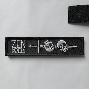以下が含まれることがあります： 「ZEN DEVILS」の文字と、ドクロと剣のデザインが施された黒色の刺繍パッチ。長方形のパッチで、裏面には面ファスナーが付いています。デザインは白い糸で表現されています。