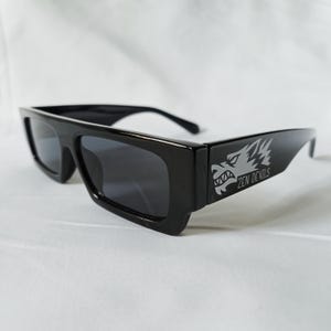 Puede incluir: Gafas de sol negras con montura rectangular gruesa. Las patillas presentan un gráfico plateado de una cabeza de lobo y el texto "ZEN DEVILS". Las lentes son oscuras, sobre un fondo blanco.