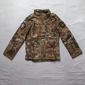 Op de afbeelding: Camouflage jas met een hoge kraag en meerdere zakken. De jas heeft een mix van bruine, groene en beige kleuren. Op beide mouwen zijn patches zichtbaar. Ontworpen voor buitenactiviteiten.