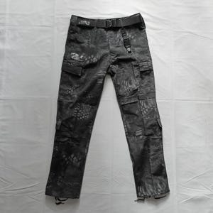 May include: Dark grey camouflage cargo trousers with multiple pockets and a black belt. The trousers feature a distinctive pattern and are made from a robust material. The belt has a metal buckle and a loop for accessories.