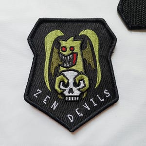 May include: Black, shield-shaped patch with the text "ZEN DEVILS". The patch features a green cartoon demon holding a white skull. The demon has red eyes and a red tongue. The patch is embroidered with black thread.