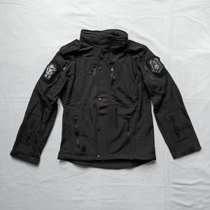 Op de afbeelding: Zwarte jas met meerdere zakken, ritsen en patches. De jas heeft een hoge kraag en knoop- en ritssluitingen. Patches met gedetailleerde ontwerpen zitten op de mouwen. De jas is gemaakt van een duurzaam, weerbestendig materiaal.