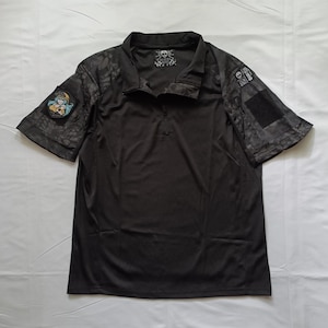 Peut inclure: Une chemise tactique noire à manches courtes avec un col zippé. Les manches présentent un motif camouflage sombre, un patch rectangulaire noir auto-agrippant et un écusson circulaire représentant un personnage aux cheveux bleus. Logo crâne au col.