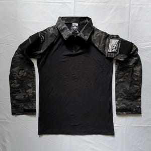 May include: Black long-sleeve shirt with a camouflage pattern on the sleeves and shoulders. The shirt has a zipper closure and a patch on the right sleeve that says "Skull Face".