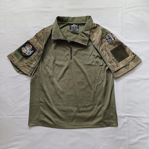 May include: An olive green short-sleeve tactical shirt with a quarter-zip collar. The sleeves feature a camouflage pattern, a circular anime-style character patch on the right, and a rectangular patch with the text ZEN DEVIL on the left.