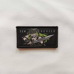 May include: A rectangular embroidered patch with a black background featuring a green-haired character wearing tactical gear and night vision goggles, holding a silver sword. The text ZEN DEVILS is printed in white capital letters above the figure.