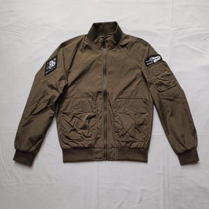 May include: Olive green bomber jacket with a zip-up front, two front pockets, and two patches on the sleeves. The jacket has a ribbed collar, cuffs, and hem. The patches feature graphic designs and text.