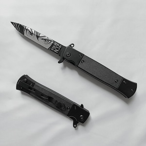 May include: Black folding knife with a textured handle and a blade featuring an anime-style eye design and the text "ZEN DE VILS". The knife is shown open and closed.