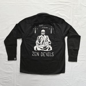 Könnte beinhalten: Schwarzes Langarmhemd mit Knöpfen und einem weißen Grafikdruck eines Skeletts, das in meditativer Haltung mit einer Katze sitzt. Der Text "DEATH IS ILLUSION" und "ZEN DEVILS" ist auf der Rückseite des Hemdes aufgedruckt.