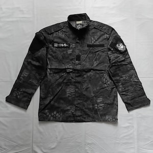 Op de afbeelding: Zwart camouflagejack met lange mouwen. De jas heeft meerdere zakken, patches en de tekst "ZEN RUI" op een patch. Het heeft een knoopsluiting aan de voorkant en een kraag. Het camouflagepatroon is een mix van donkergrijs en zwart.