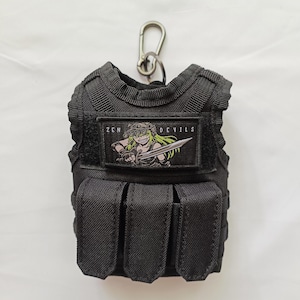 May include: A black tactical vest keychain featuring three front pouches and a rectangular patch with the text ZEN DEVILS and an illustration of a green-haired character holding a sword. The vest includes a metal carabiner clip at the top.
