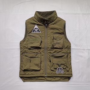 May include: Olive green quilted vest with multiple pockets and a high collar. Features a triangular patch with the text "ZEN DEVILS" and a gas mask patch. The vest has a full-length zip.