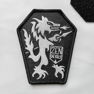 Puede incluir: Parche bordado en blanco y negro con una criatura parecida a un lobo sosteniendo una lanza. El parche tiene un borde negro y el texto "ZEN DEVILS" en un diseño de escudo. El parche está sobre un fondo blanco.