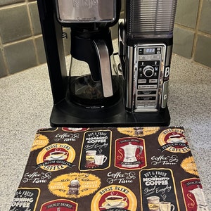 Puede incluir: Una cafetera negra Ninja con una jarra de vidrio se encuentra sobre una alfombra de cocina de temática de café marrón y roja. La cafetera tiene una pantalla digital que muestra la hora 8:33. La alfombra tiene una variedad de diseños de temática de café, incluyendo granos de café, tazas de café y cafeteras.