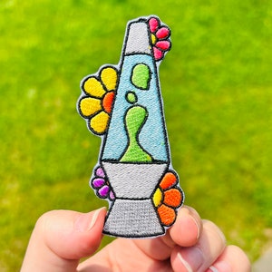 May include: A colorful embroidered iron-on patch featuring a lava lamp with a green and blue liquid inside. The lamp is surrounded by four colorful flowers.