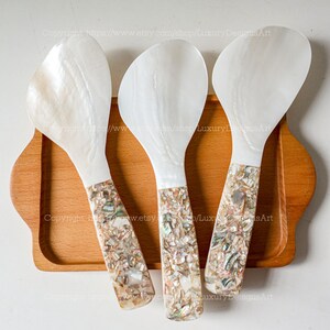 May include: Three white mother of pearl serving spoons with iridescent shell inlay handles. The spoons are arranged on a wooden tray.