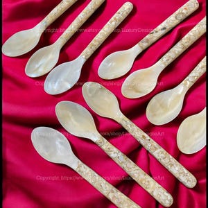 May include: A set of ten mother of pearl spoons with a natural shell inlay design. The spoons are arranged on a red fabric background.
