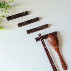 May include: A set of dark brown wooden chopsticks and a matching spoon resting on a dark brown wooden chopstick holder. The chopsticks and spoon are made of dark brown wood and have a smooth finish.