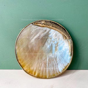 Könnte beinhalten: Eine runde, schillernde Perlmutt-Schale mit einem goldfarbenen Fischdesign. Die Schale ist perfekt für die Aufbewahrung von kleinen Gegenständen wie Schmuck oder Schlüssel.