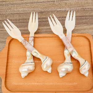 Op de afbeelding: Vier decoratieve vorken met een schelpmotief. De vorken hebben witte tanden en handvatten met een parelmoer, gemarmerd patroon. De handvatten zijn gevormd als schelpen en liggen op een houten dienblad.