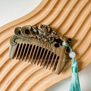 K&ouml;nnte beinhalten: Ein dunkelbrauner Holzkamm mit aufwendigen floralen Schnitzereien und einem gr&uuml;nen Quasten. Der Kamm hat eine jadegr&uuml;ne Perle an der Quaste.