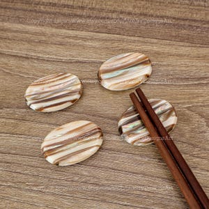 Op de afbeelding: Vier ovale, iriserende eetstokjes rusten op een houten oppervlak. Eén rust heeft een paar bruine eetstokjes erop liggen.