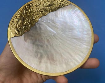 Puede incluir: Plato decorativo redondo con centro de nácar y borde de metal dorado. El borde superior presenta un diseño de relieve dorado adornado con peces y otros elementos acuáticos. El plato se sostiene en una mano sobre un fondo azul.