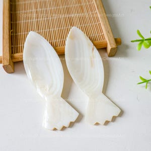 May include: Two white mother of pearl fish shaped spoons. The spoons are on a white surface with a bamboo mat in the background.