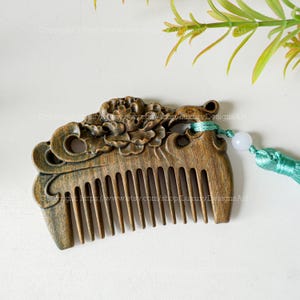 K&ouml;nnte beinhalten: Ein Holzkamm mit einem geschnitzten Blumendesign und einem gr&uuml;nen Quasten. Der Kamm ist dunkelbraun und hat eine glatte Oberfl&auml;che. Die Quaste ist aus einem weichen, seidenartigen Material.