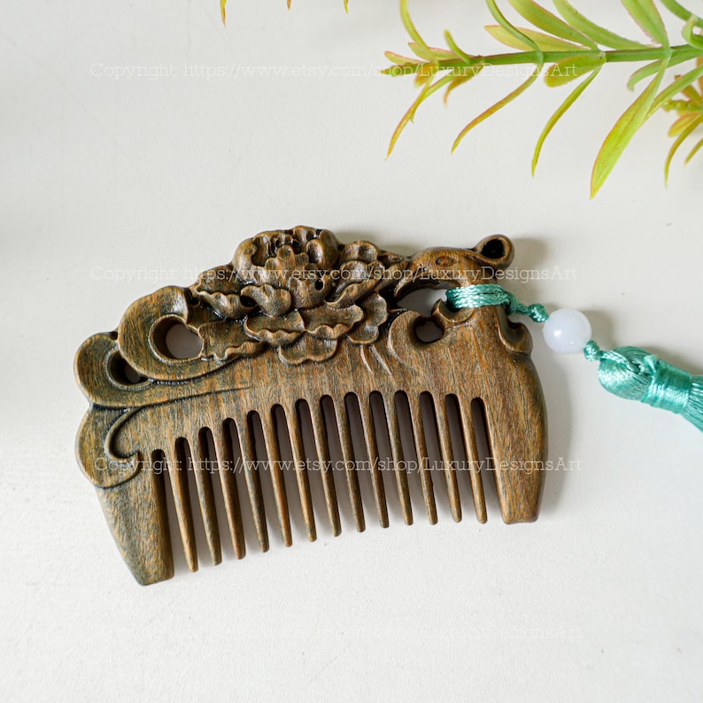 K&ouml;nnte beinhalten: Ein Holzkamm mit einem geschnitzten Blumendesign und einem gr&uuml;nen Quasten. Der Kamm ist dunkelbraun und hat eine glatte Oberfl&auml;che. Die Quaste ist aus einem weichen, seidenartigen Material.