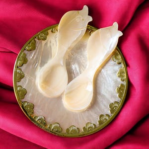 Puede incluir: Dos cucharas blancas de nácar con forma de pez, descansando sobre un plato redondo de nácar blanco con un borde dorado y un diseño floral.