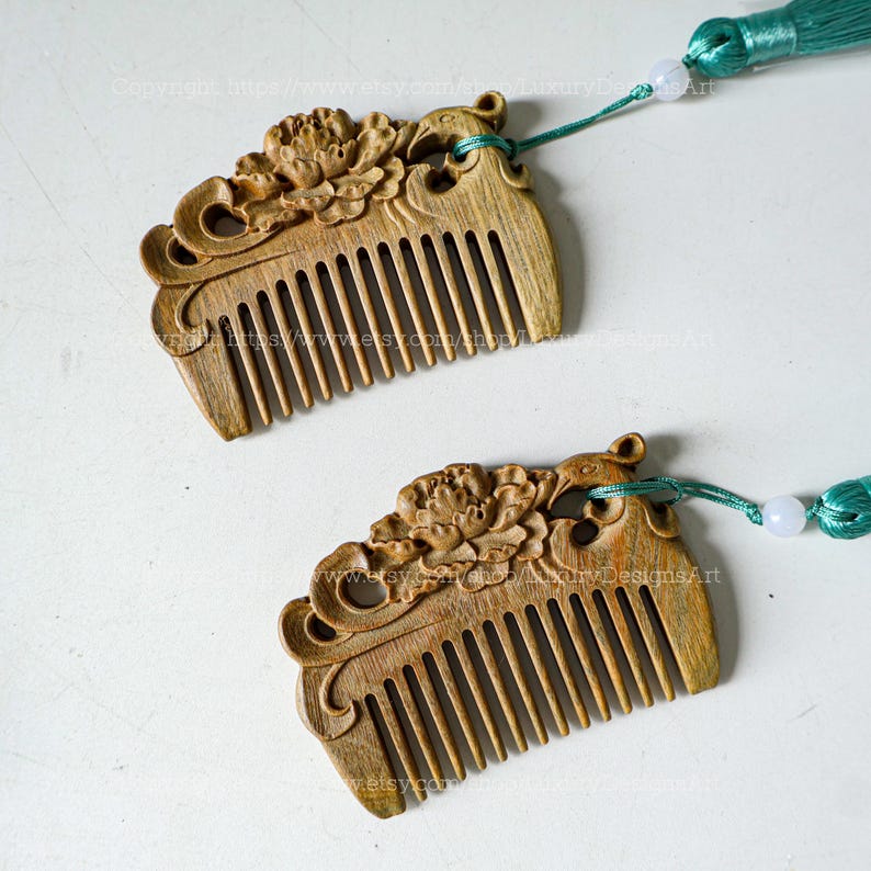 K&ouml;nnte beinhalten: Zwei kunstvoll geschnitzte Holzk&auml;mme mit Blumendesigns und gr&uuml;nen Quasten. Die K&auml;mme haben eine nat&uuml;rliche Holzoptik und sind mit einer wei&szlig;en Perle zusammengebunden.