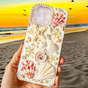 May include: A white phone case decorated with seashells, pearls, and starfish. The case has a pink and white seashell, a large white seashell, and a white starfish in the center. The case is surrounded by smaller seashells and pearls.