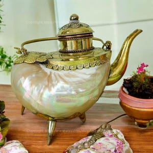 May include: A gold and brass teapot with a shell-shaped body and three legs. The teapot has a lid with intricate details and a spout on the side. The teapot is sitting on a wooden surface.