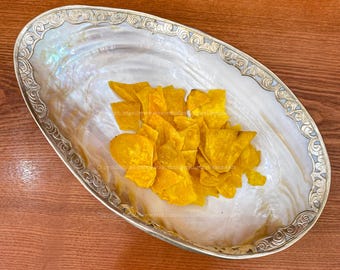 May include: A white oval serving dish with a silver rim and intricate designs. The dish is filled with a pile of yellow tortilla chips.
