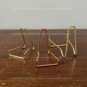 Puede incluir: Tres expositores metálicos en tonos dorados y cobrizos. Los expositores están diseñados para sujetar objetos pequeños. Los expositores están sobre una superficie de madera. El texto "Copyright https://www.etsy.com/shop/LuxuryDesignsArt" es visible.