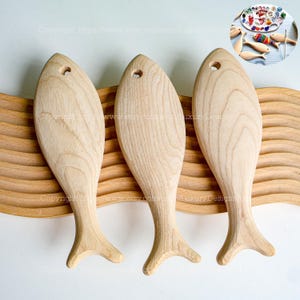 Op de afbeelding: Drie onafgewerkte houten visvormen, elk met een klein gat geboord in de bovenkant, zijn gerangschikt op een houten oppervlak met een golvend patroon. De vissen zijn allemaal in dezelfde richting gericht, met hun staarten naar rechts.