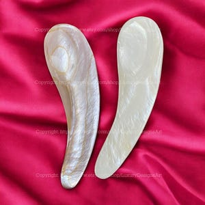 Puede incluir: Dos herramientas de belleza curvas y nacaradas con un acabado liso y pulido. Una tiene un brillo sutil e iridiscente con tonos dorados, mientras que la otra es de color blanco crema. Descansan sobre una tela roja intensa.