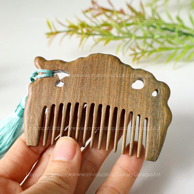K&ouml;nnte beinhalten: Ein Holzkamm mit einem geschnitzten Design und einem t&uuml;rkisfarbenen Quasten. Der Kamm hat eine nat&uuml;rliche Holzoptik und besteht aus einem einzigen St&uuml;ck Holz.