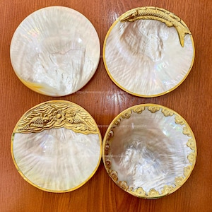 May include: Four round, iridescent mother-of-pearl dishes with gold trim. Two have gold fish designs, one a floral design, and one a cloud design. The dishes are on a brown wooden surface.