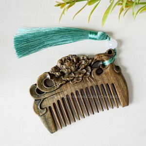K&ouml;nnte beinhalten: Ein Holzkamm mit einem geschnitzten Blumenmuster und einem gr&uuml;nen Quasten. Der Kamm hat eine dunkelbraune Oberfl&auml;che und ist mit einer geschnitzten Blume verziert.
