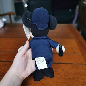 K&ouml;nnte beinhalten: Ein Pl&uuml;sch-Mickey-Mouse-Spielzeug, das einen blauen Hut und eine blaue Jacke tr&auml;gt. Das Spielzeug h&auml;lt einen schwarz-wei&szlig;en Stock in seiner rechten Hand. Das Spielzeug steht auf einer Holzoberfl&auml;che.